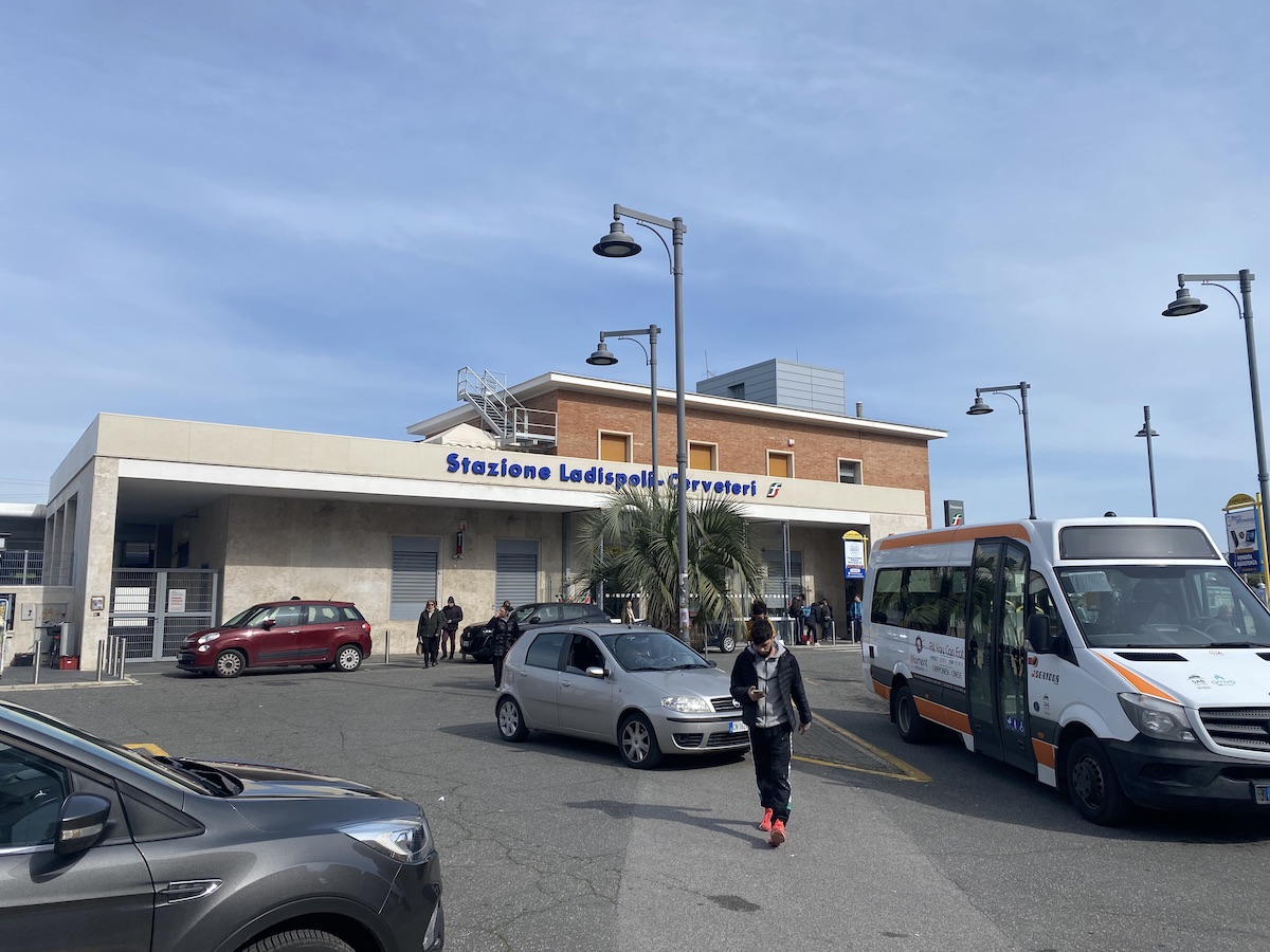 A view of the train station with pedestrians looking at their phones and several parked vehicles.