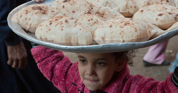 In Cairo, Egypt, a young child wearing a pink jacket carries a battered alumium tray on their head loaded with freshly baked flatbreads. 