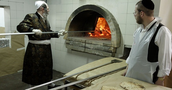 Orthdox Jews baking unleavened bread, matzo, in a wood-fired oven. On the left a man in a long frock coat wearing a white skullcap is manipulating the long aluminium handle of a peel for managing the breads in the oven. On the right a younger man wearing a white plastic disposable apron looks on.