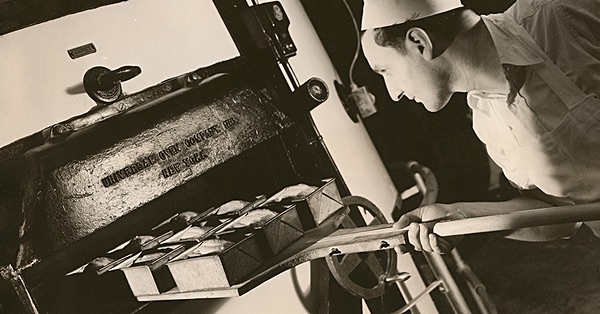 Monochrome image of a baker putting loaf tins into an oven. The baker wears a pristine white chef's hat and is leaning forward on the right. The oven door on the left is open to receive the tins, which are lined up on a wooden peel.