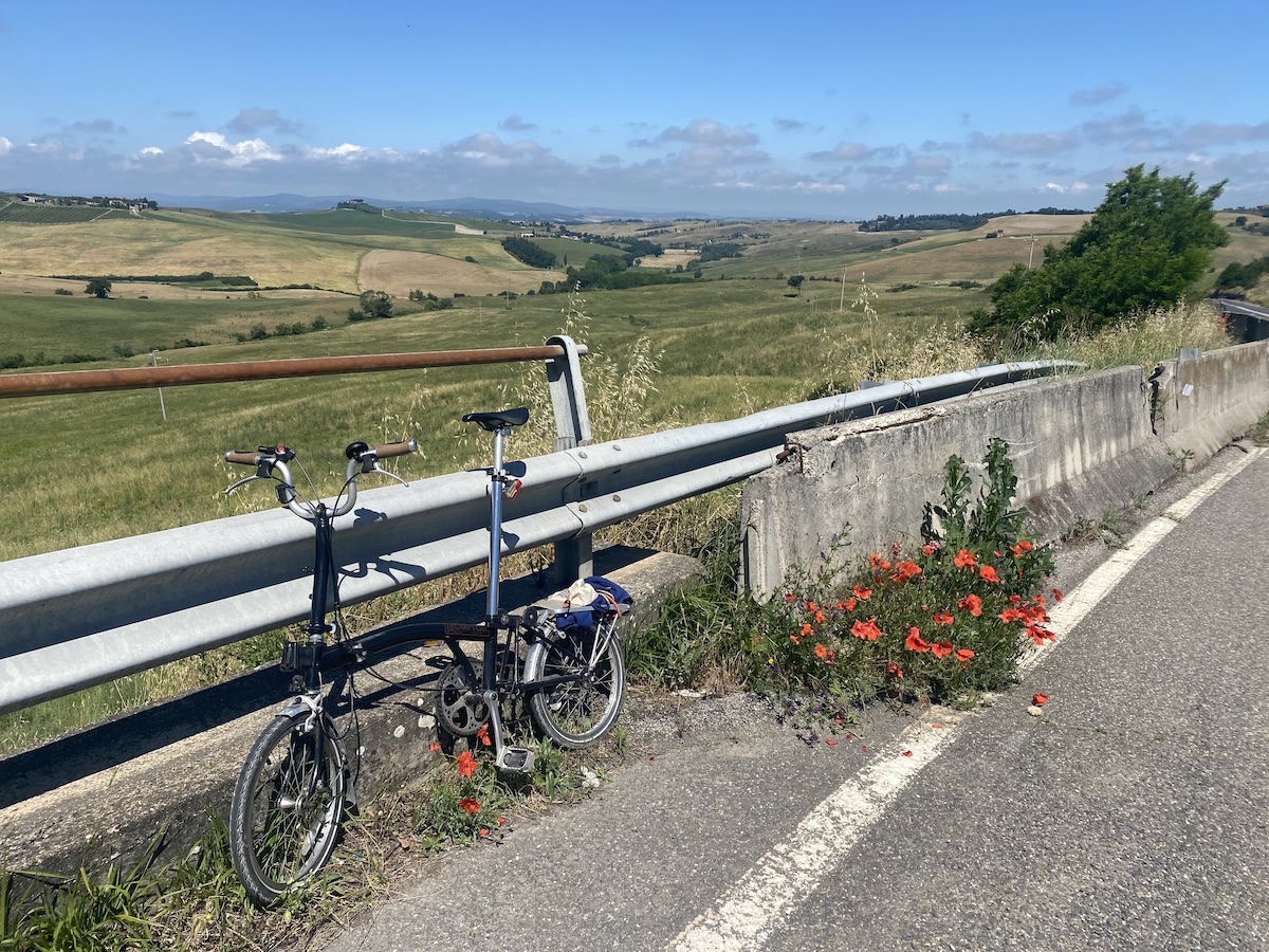 Brompton bicycle leaning against road guardrail with a patch of bright red poppies behind it. In the distance the town of Buonconvento in a landscape of Tuscan hills.