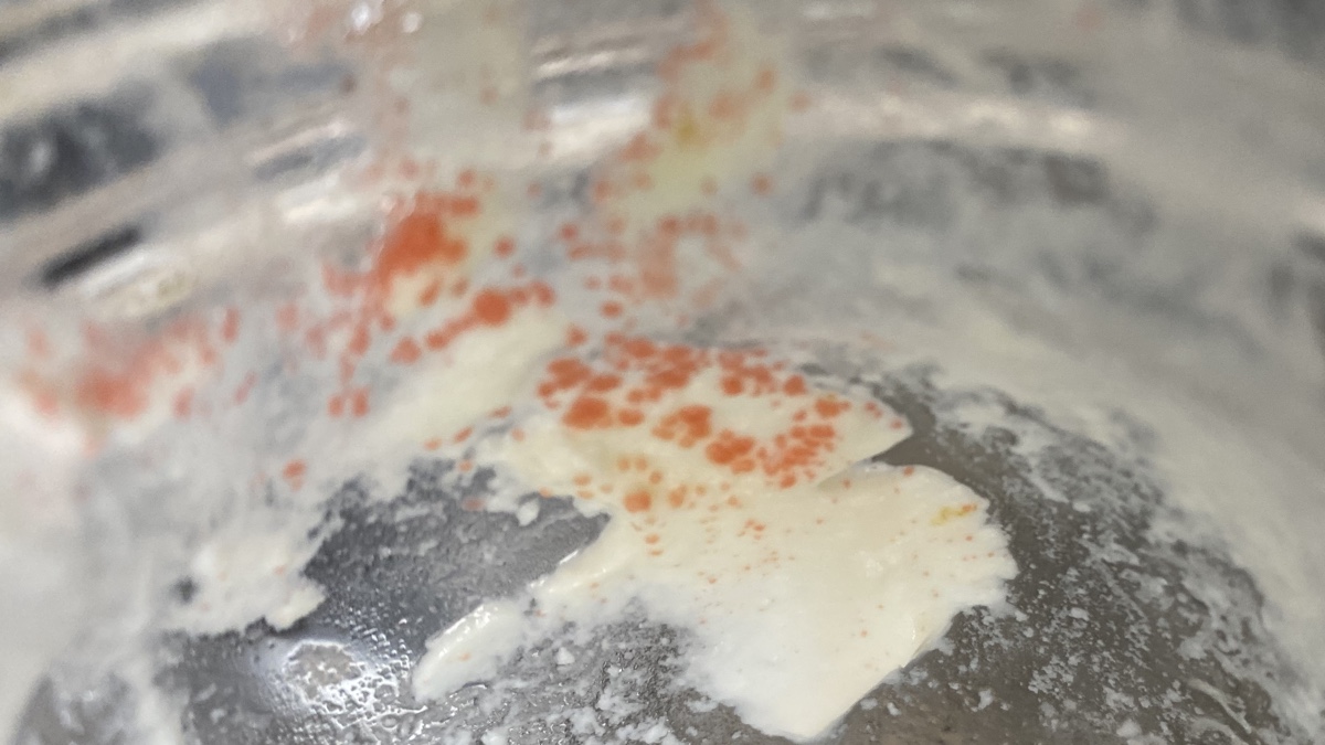 Spots of a reddish-orange mould growing on off-white yoghurt on the side of a glass jar.