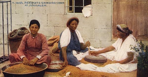 Three women seated on the ground in front of a building. On the left, a woman in a red dress has her hands in a bowl of wheat seeds. On the right, a woman in a blue dress holds the handle of a rotary quern. On the far right, a woman in white feeds wheat into the hole in the centre of the quern. From a 1912 postcard entitled Peasants Grinding Corn at Jerusalem.