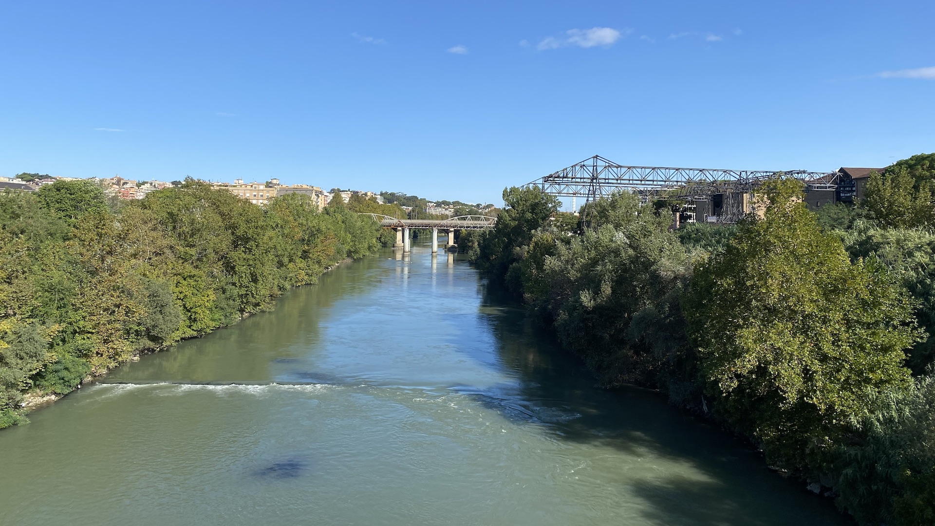 Looking down on the Tiber river. In the river there is a small weir with white water tumbling over it. The river banks are green, and there are the outline of an older bridge and the industrial remains of the old gasometer and unloading conveyor.